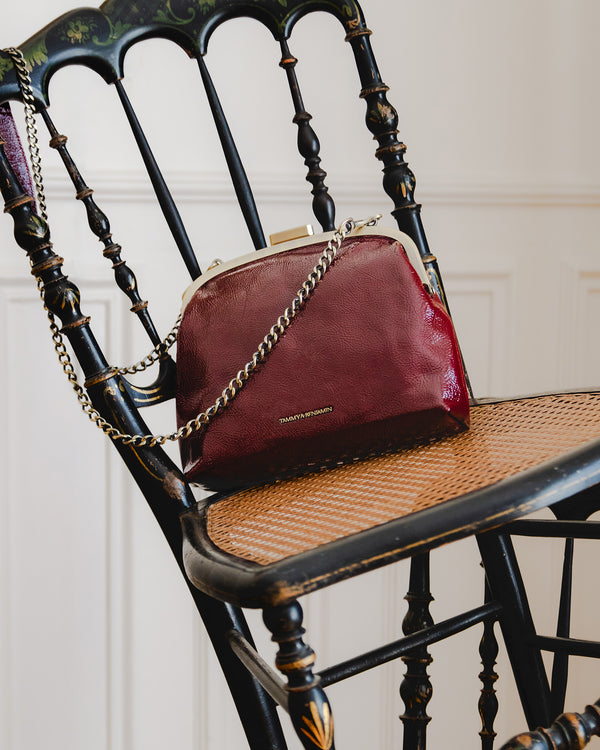 EMMA bag in burgundy naplak, front view, resting on a chair, gold clasp, chain strap draped across
