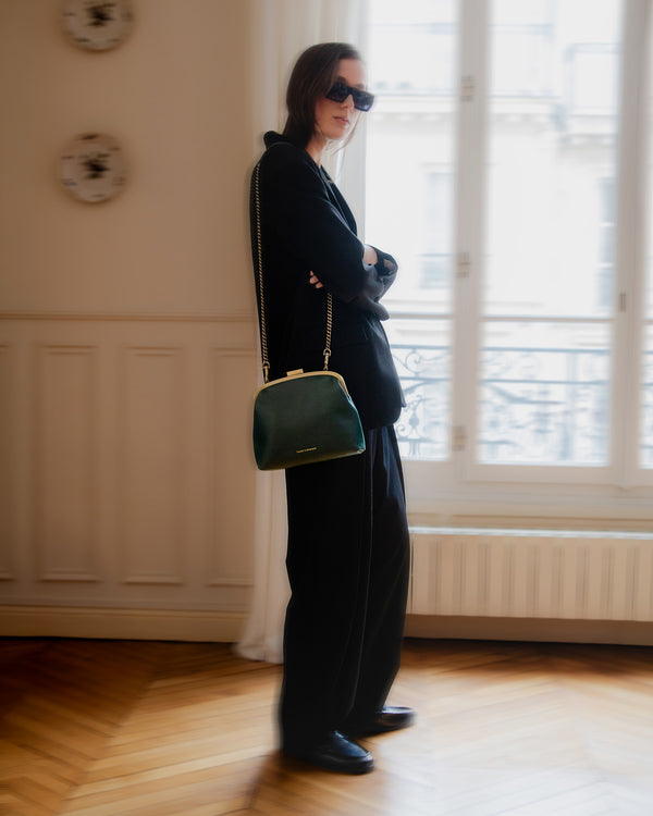 Sac vert émeraude porté en bandoulière avec chaîne longue, femme de profil près d'une fenêtre