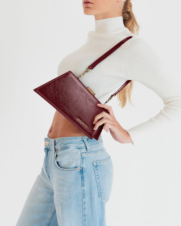 JAY bag in Bordeaux naplak, held in hand with shoulder strap, side view over jeans