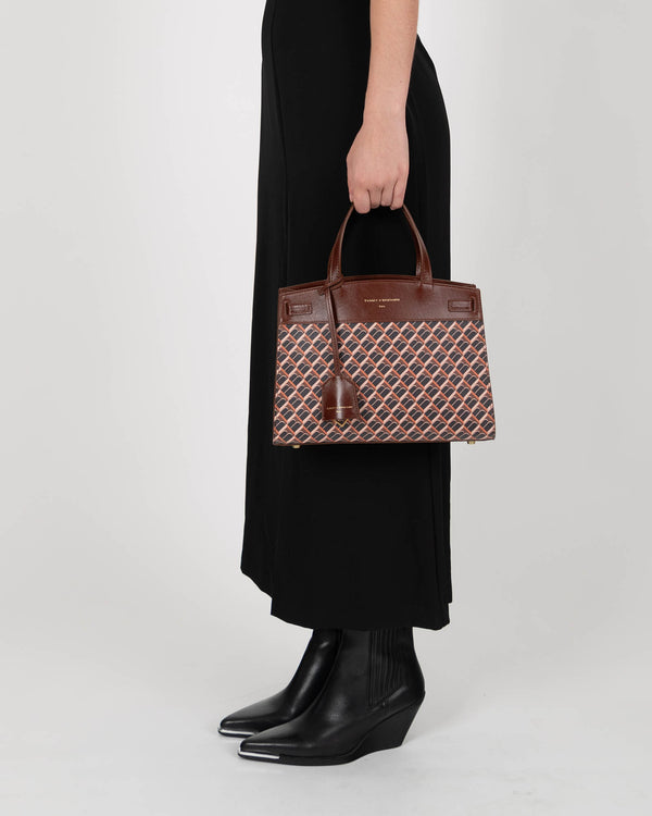 Sac MADISON brun monogram porté à la main par une femme en robe noire, vue de profil