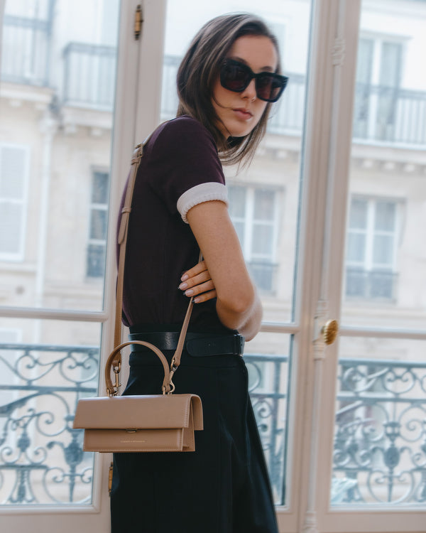 Sac THEA MINI beige porté en bandoulière, vue de profil dans l'embrasure d'une fenêtre parisienne