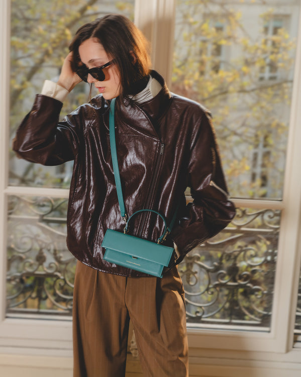 Sac THEA MINI Turquoise porté en bandoulière, poitrine, veste cuir marron, fenêtre parisienne