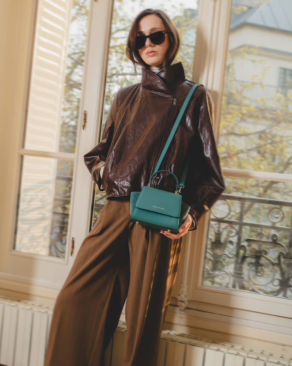 ALEXIA NANO Turquoise, porté en bandoulière à la main, devant une fenêtre
