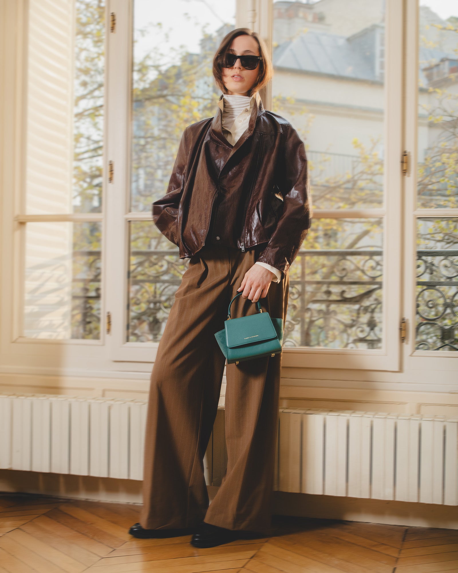Sac ALEXIA NANO turquoise porté à la main par une femme en tenue brune, devant une fenêtre parisienne