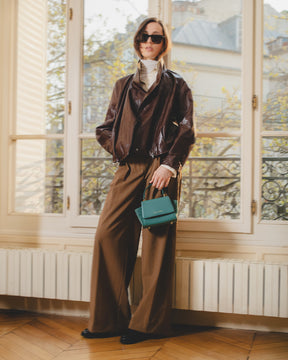 Sac ALEXIA NANO turquoise porté à la main par une femme en tenue brune, devant une fenêtre parisienne