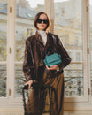 Sac ALEXIA NANO turquoise tenu à la main, anse courte, porté par une femme en veste en cuir