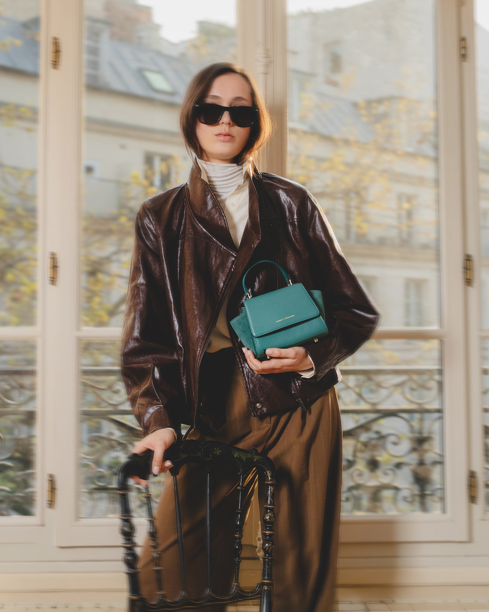 Sac ALEXIA NANO turquoise tenu à la main, anse courte, porté par une femme en veste en cuir