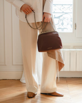 Sac EMMA brun tenu à la main par la chaîne, porté par une femme en pantalon à rayures
