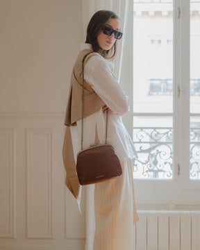 Sac EMMA brun porté à l'épaule avec bandoulière chaîne, vue de côté près d'une fenêtre