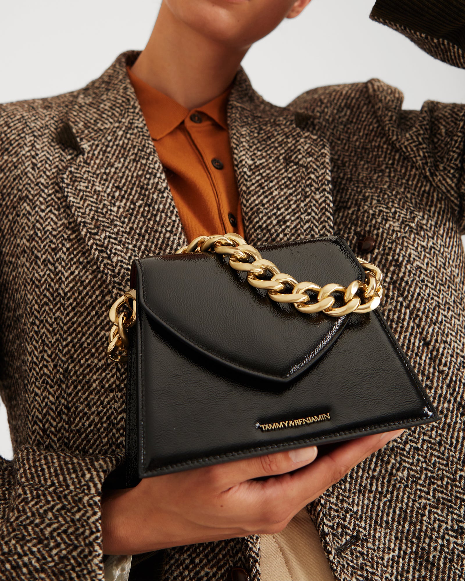 GABRIELLE bag in black naplak leather held in both hands, pointed flap and chunky gold chain handle over herringbone jacket