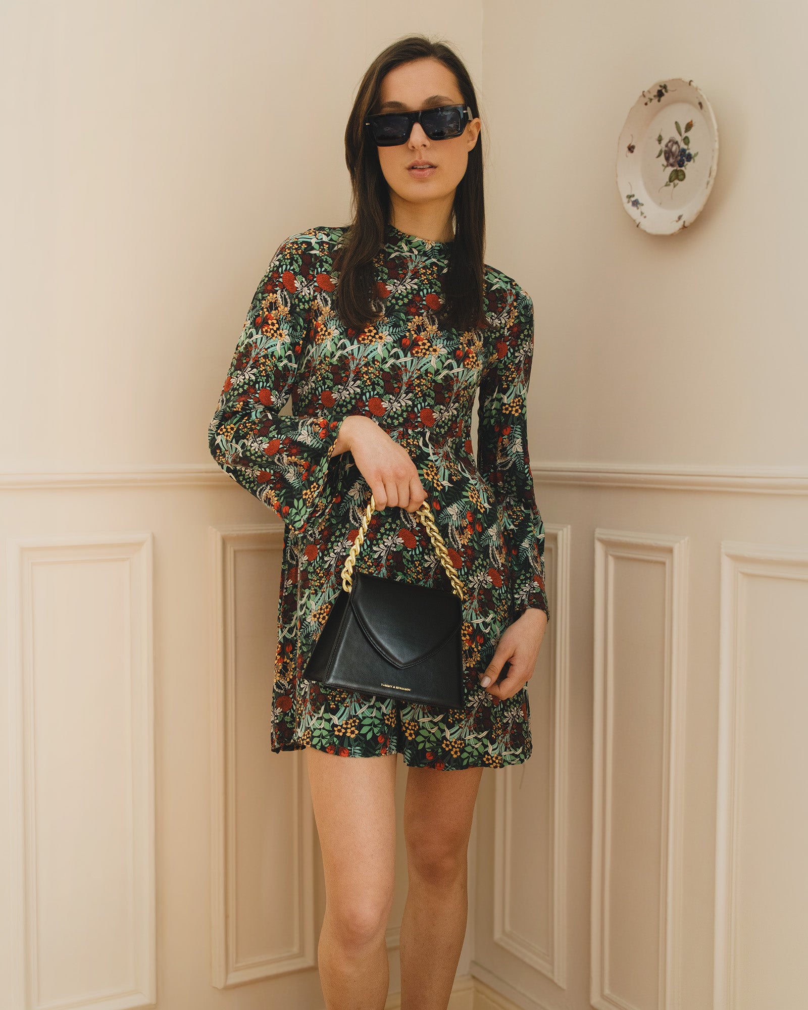 Sac GABRIELLE noir porté à la main par sa chaîne dorée, de face, avec robe fleurie