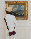 Sac JEANNE BAGUETTE Mahogany porté en bandoulière, vu de dos, devant un tableau dans un intérieur chic