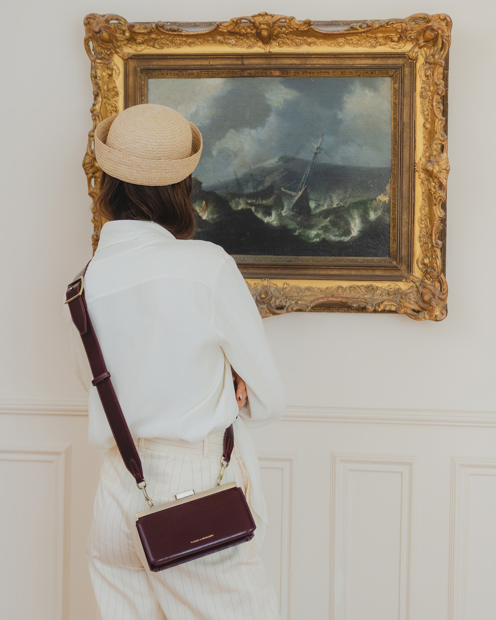 Sac JEANNE BAGUETTE Mahogany porté en bandoulière, vu de dos, devant un tableau dans un intérieur chic