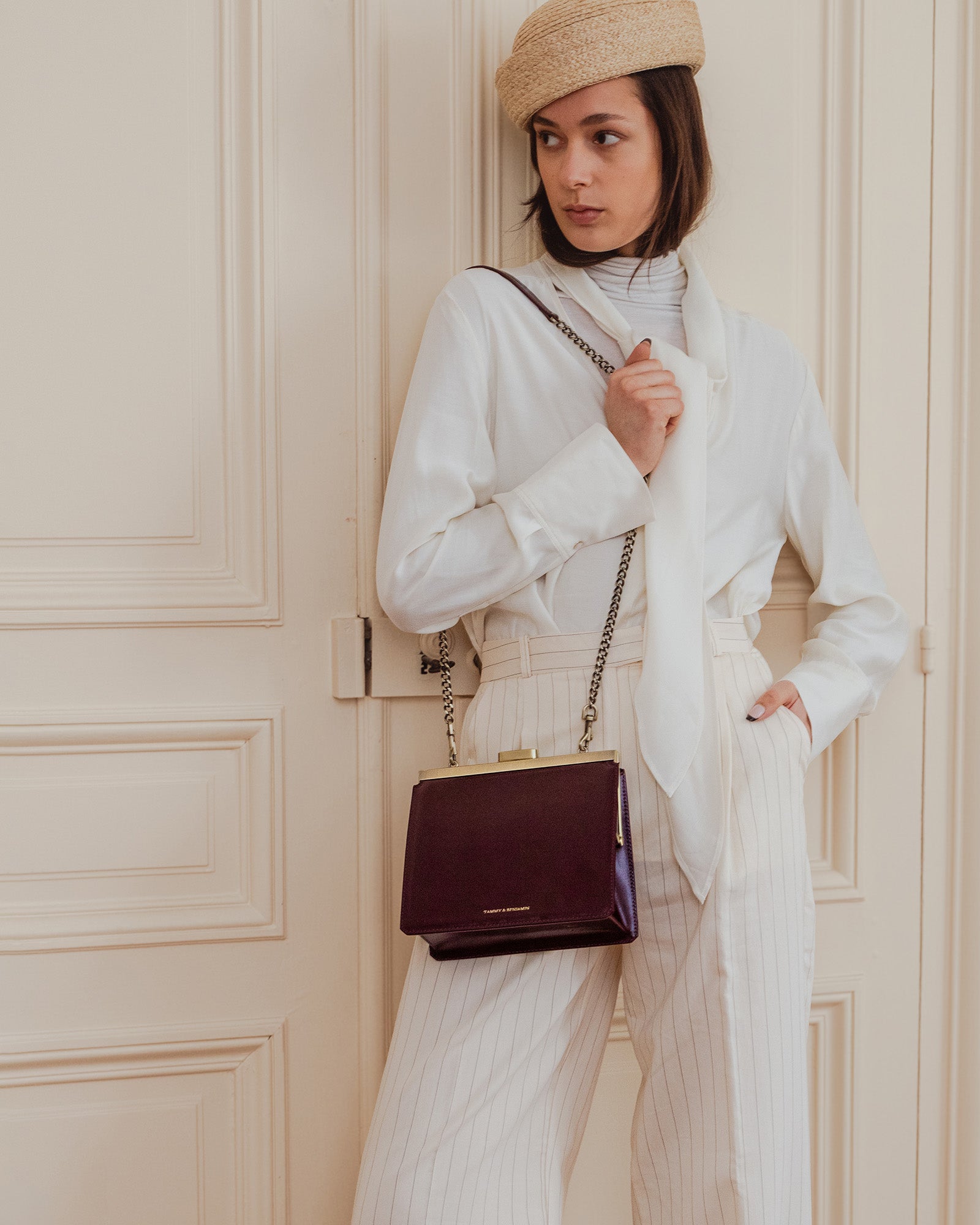 Sac JEANNE Mahogany porté en bandoulière avec chaîne, posé contre une porte, vue de face
