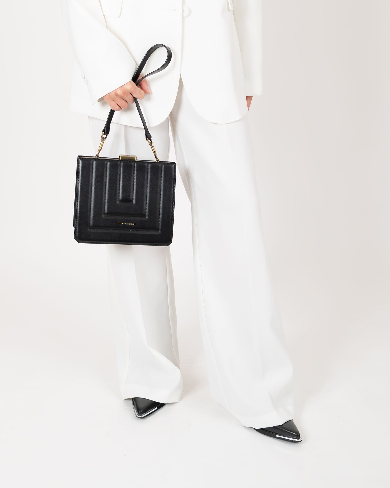 Sac JUDE noir matelassé tenu à la main par l'anse, porté par une femme en tailleur blanc