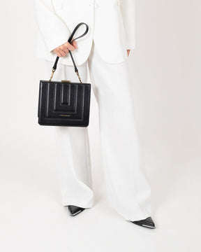 Sac JUDE noir matelassé tenu à la main par l'anse, porté par une femme en tailleur blanc