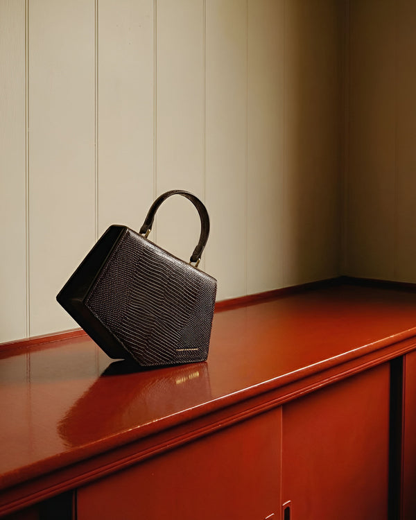 Sac PENTAGONE brun embossé lézard, vue de côté, poignée courte, posé sur un meuble laqué rouge