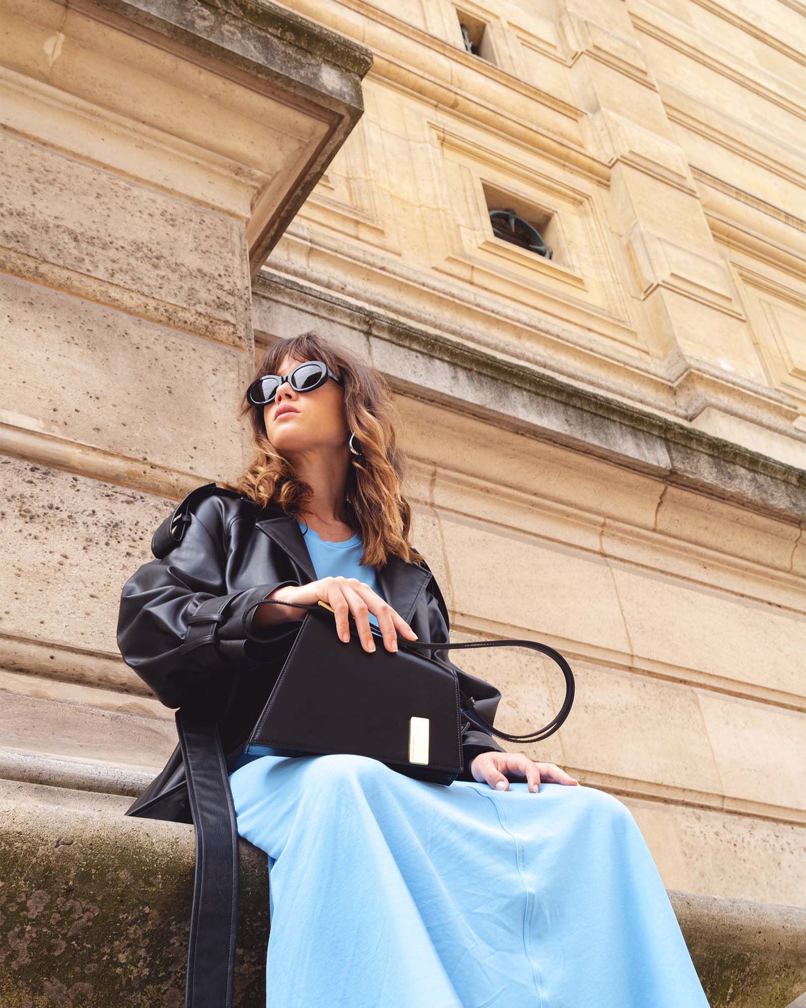 Sac TARA noir porté à la main, silhouette graphique, assise en extérieur devant un bâtiment