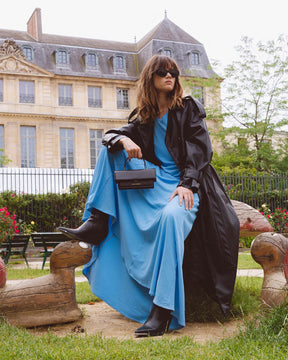 Sac THEA MINI noir tenu à la main par une femme assise en robe bleue et trench noir