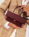 THEA MINI bag in Mahogany held by its top handle, gold clasp, carried in front of a check blazer