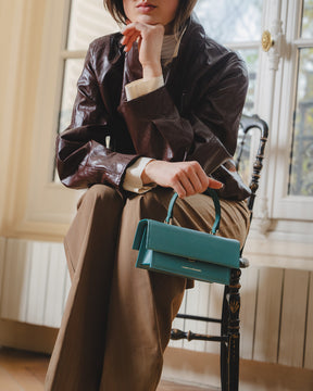 THEA MINI bag in turquoise, held by its handle, carried by a seated woman