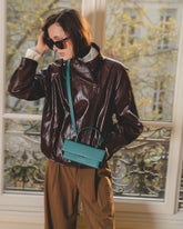 THEA MINI bag in Turquoise worn crossbody, brown leather jacket, Parisian window background