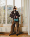 Sac THEA Turquoise porté à la main par poignée rigide, devant une fenêtre parisienne
