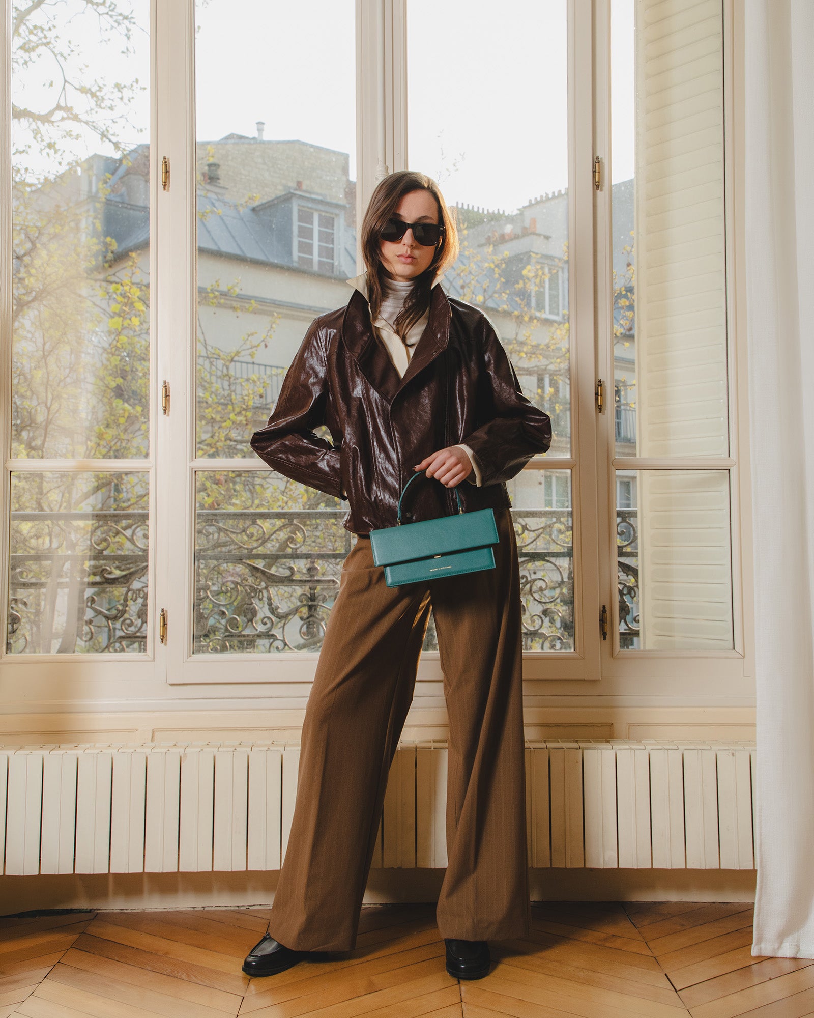 Sac THEA Turquoise porté à la main par poignée rigide, devant une fenêtre parisienne