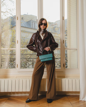 Sac THEA Turquoise porté à la main par poignée rigide, devant une fenêtre parisienne